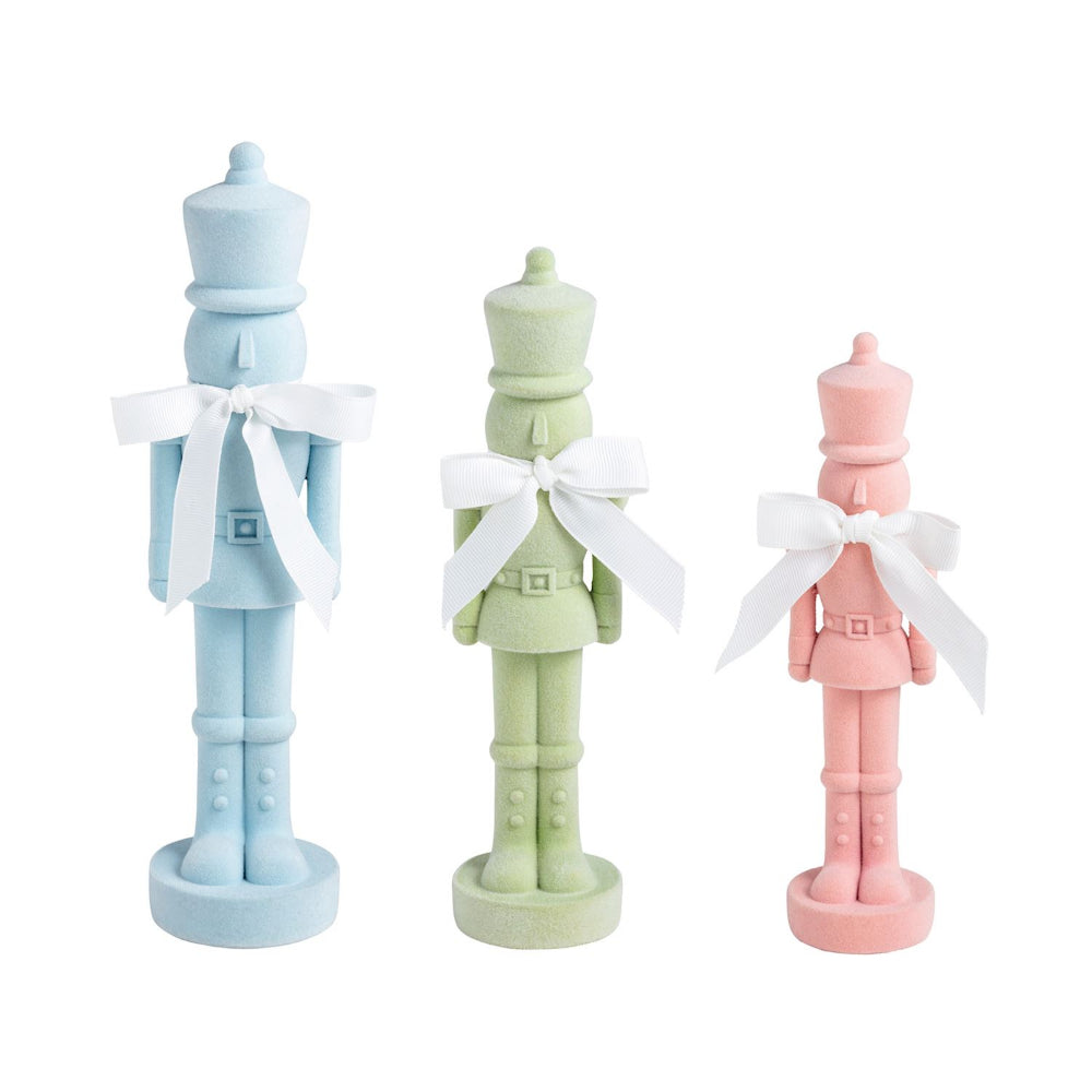 Three decorative nutcracker figurines standing side by side on a white background, each with a white ribbon bow: one light blue, one pale green, and one pale pink.
