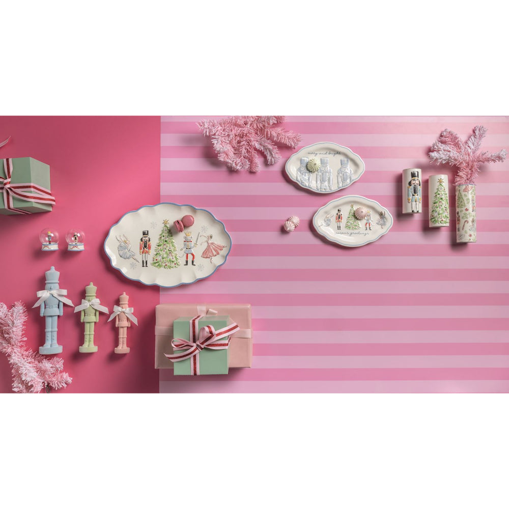 A styled flat-lay of a ceramic holiday tableware set on a pink and white striped background. The set includes two scalloped oval platters with festive illustrations, two matching cylindrical vases, and several nutcracker figurines in pastel colors, alongside wrapped gift boxes and pink decorative branches.