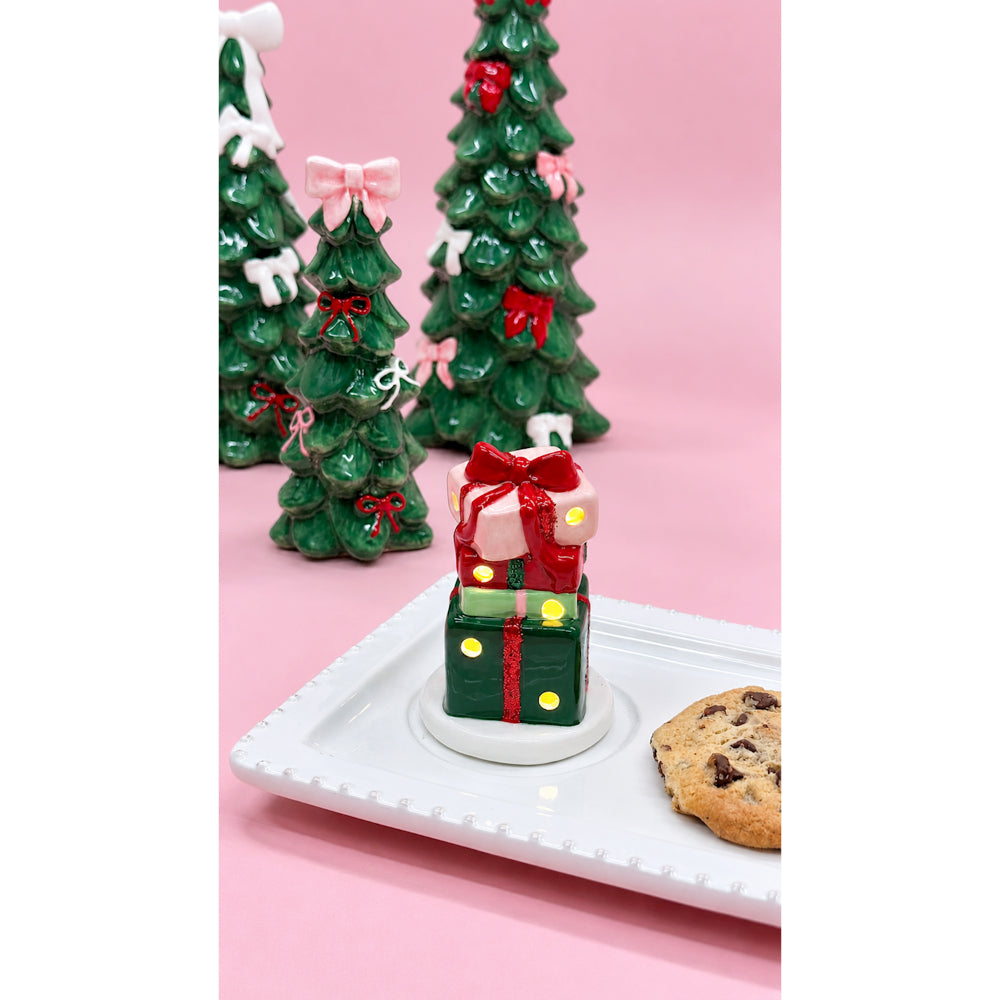 A small Christmas-themed gift box candle sits on a white square plate, styled with miniature green Christmas trees and ornaments in the background against a solid pink backdrop.