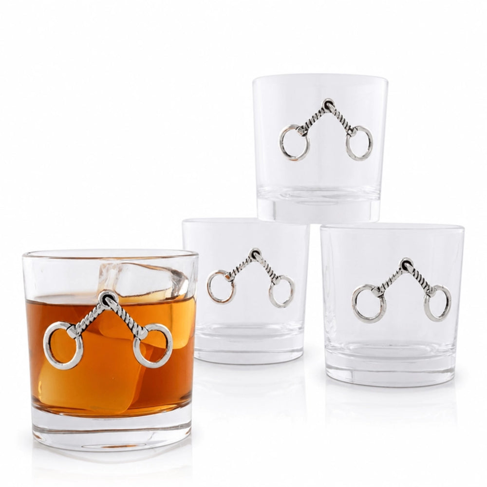 Four clear glass rocks tumblers with silver horse-bit metal accents, with one glass filled with amber liquor and an ice cube on a white background.