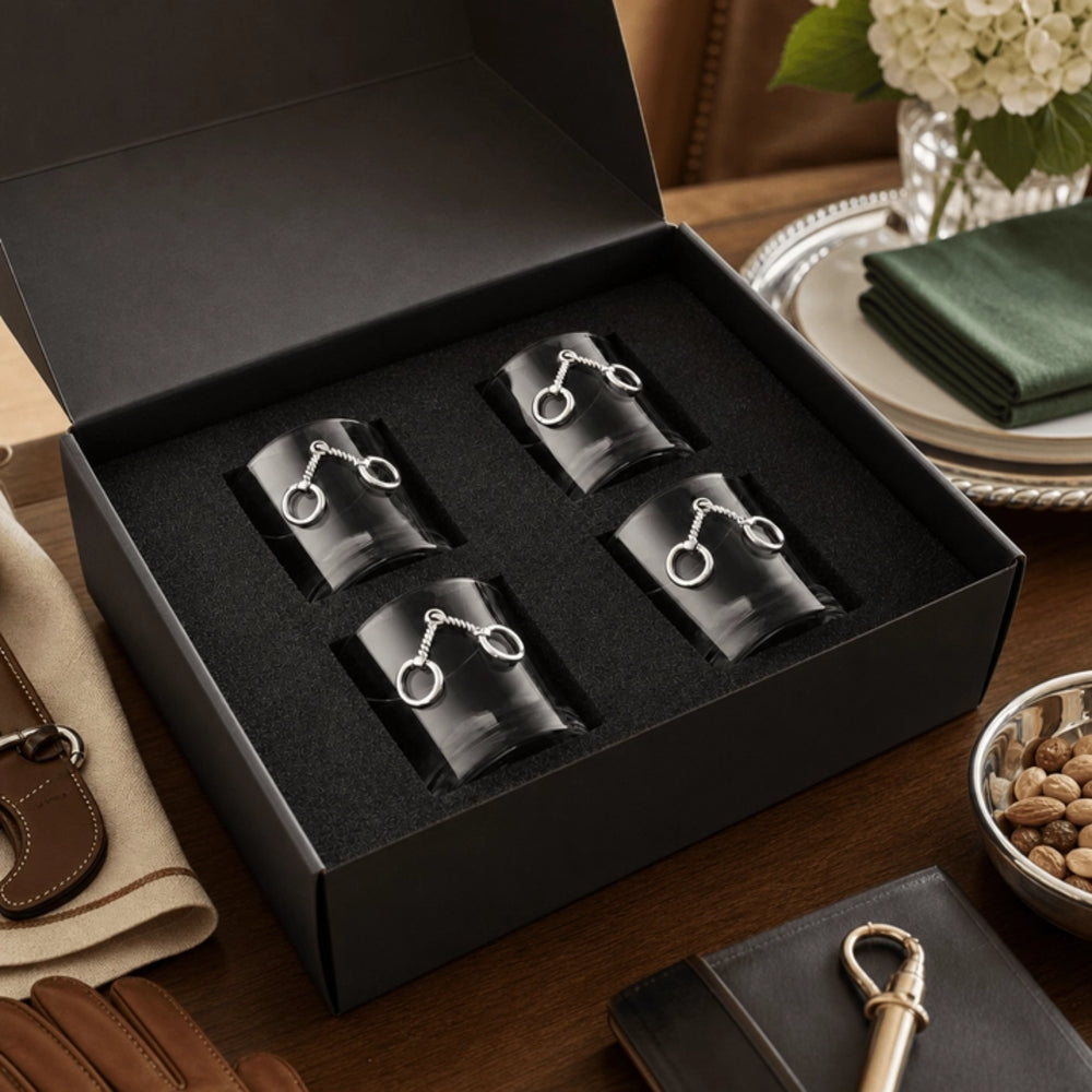 Four black cylindrical accessories with silver horsebit rings, presented in a black gift box on a wooden table with lifestyle props.