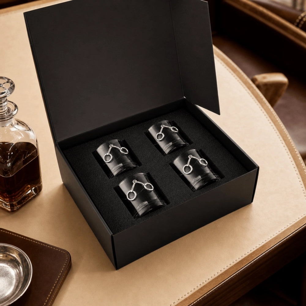 A black presentation box opened on a beige leather surface, containing four black cylindrical accessories with silver horse-bit style rings. A glass decanter and leather tray are partially visible nearby.