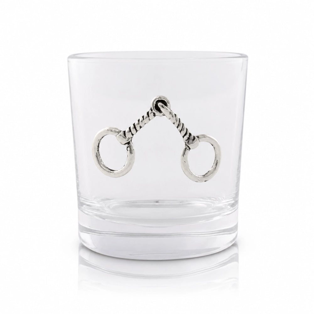 A clear glass rocks tumbler featuring a decorative silver horse bit with two rings on the front, set against a clean white background.