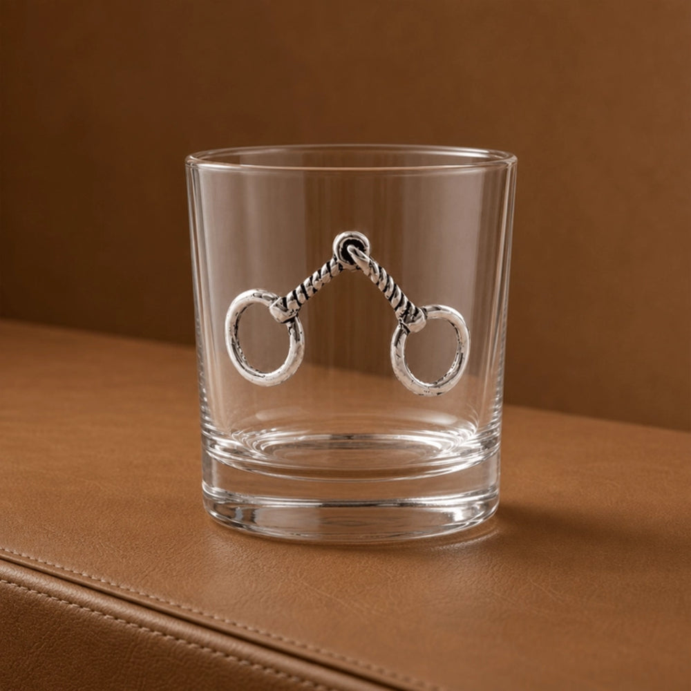 A clear lowball glass with a silver horse-bit style ornament on the front, set on a brown leather surface against a matching brown background.