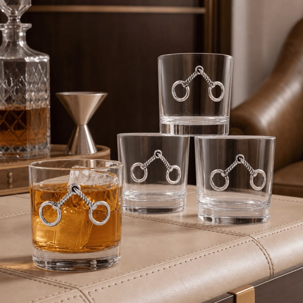 Four clear whiskey glasses with silver horsebit designs on the sides, arranged on a tan leather bar surface with a whiskey bottle and decanter in the background.