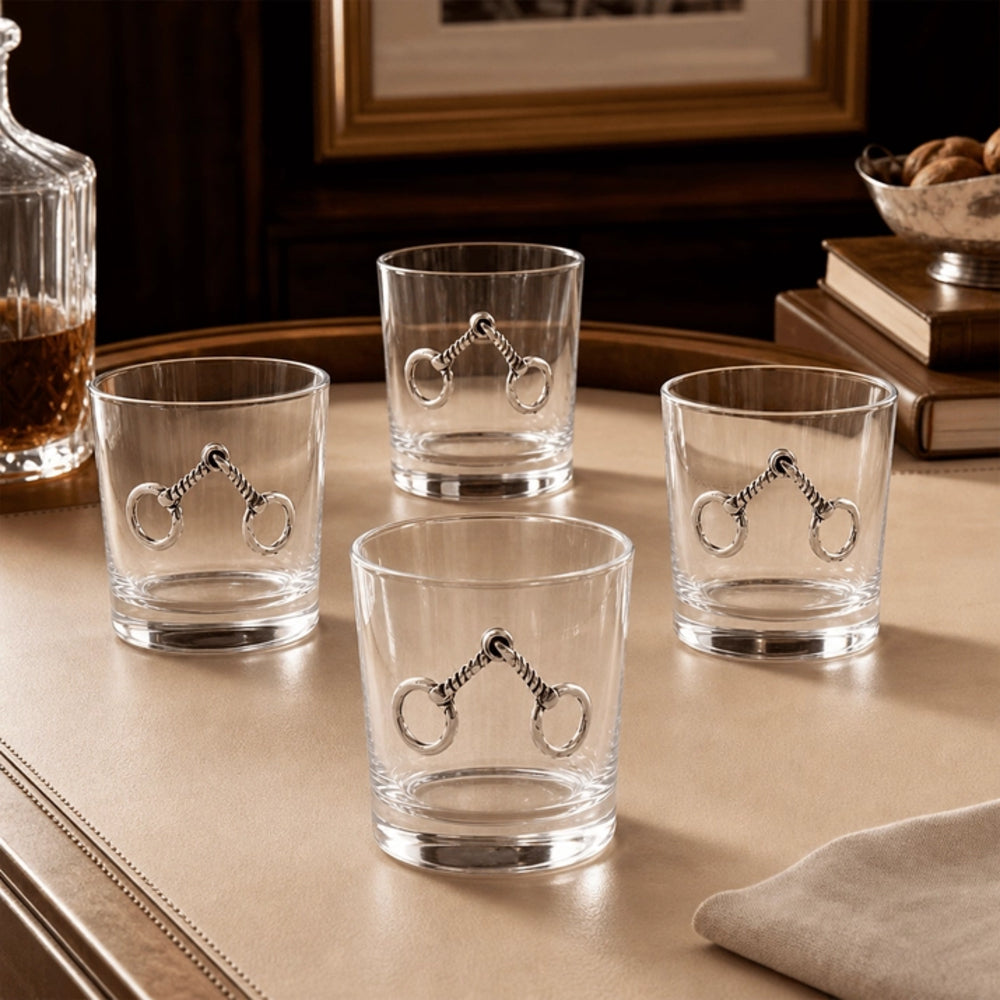Four clear glass rocks tumblers with silver horsebit designs on the front, arranged on a beige table with a bottle, books, and framed picture in the background.