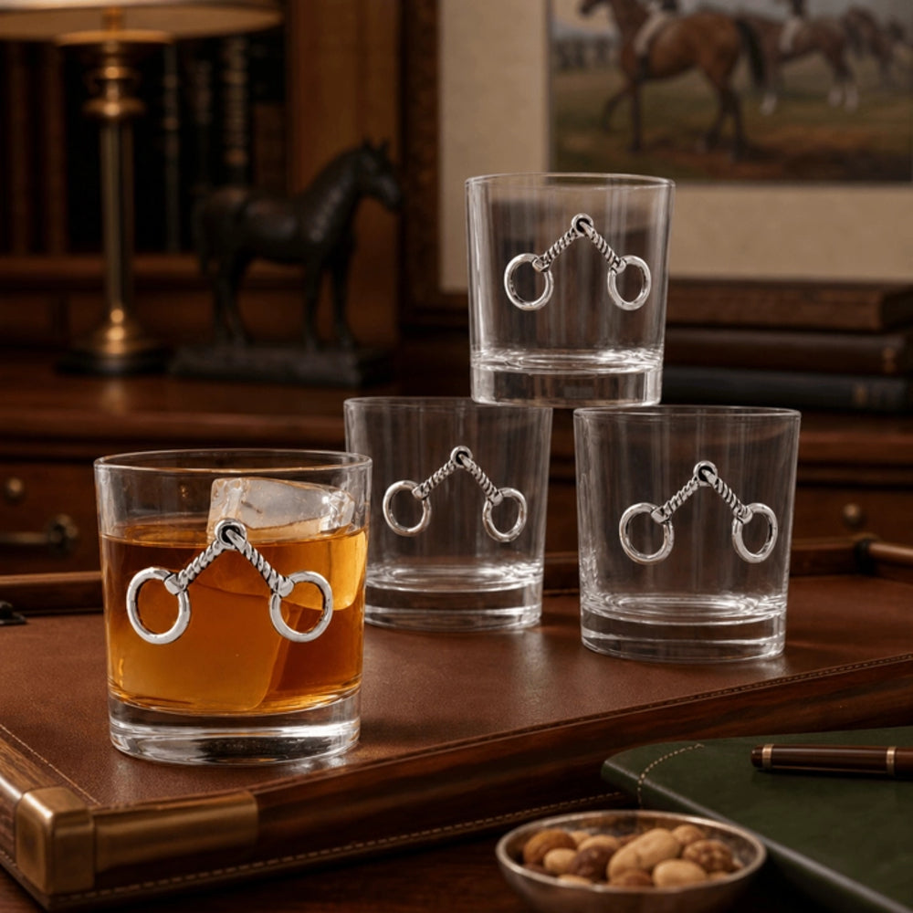 Three clear rocks glasses with silver horse-bit ring charms, with one glass filled with amber whiskey, displayed on a leather tray in a warm, rustic interior setting.