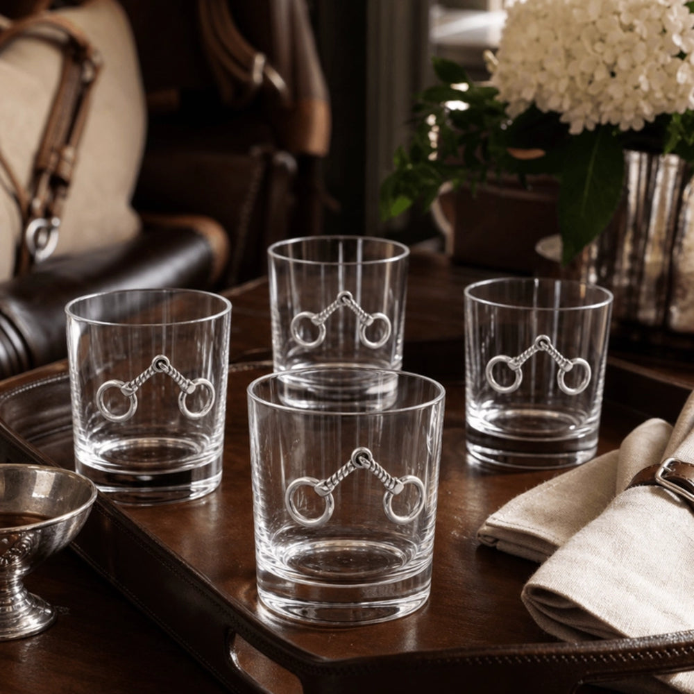 Four clear glass rocks tumblers with an engraved horse bit design, arranged on a dark wooden tray in a styled interior setting.