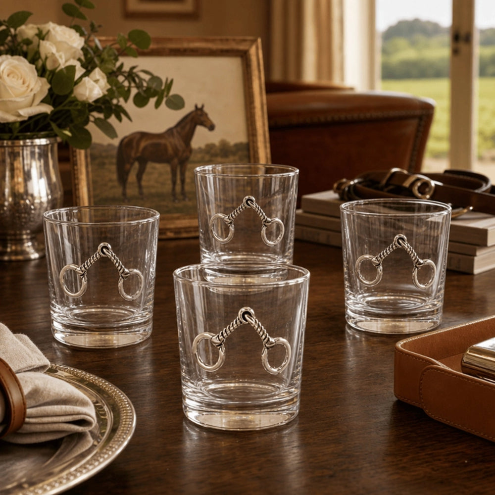 Four clear glass tumblers with silver horse-bit designs, arranged on a dark wooden table in a styled interior setting with books, a leather box, and a framed picture of a horse.