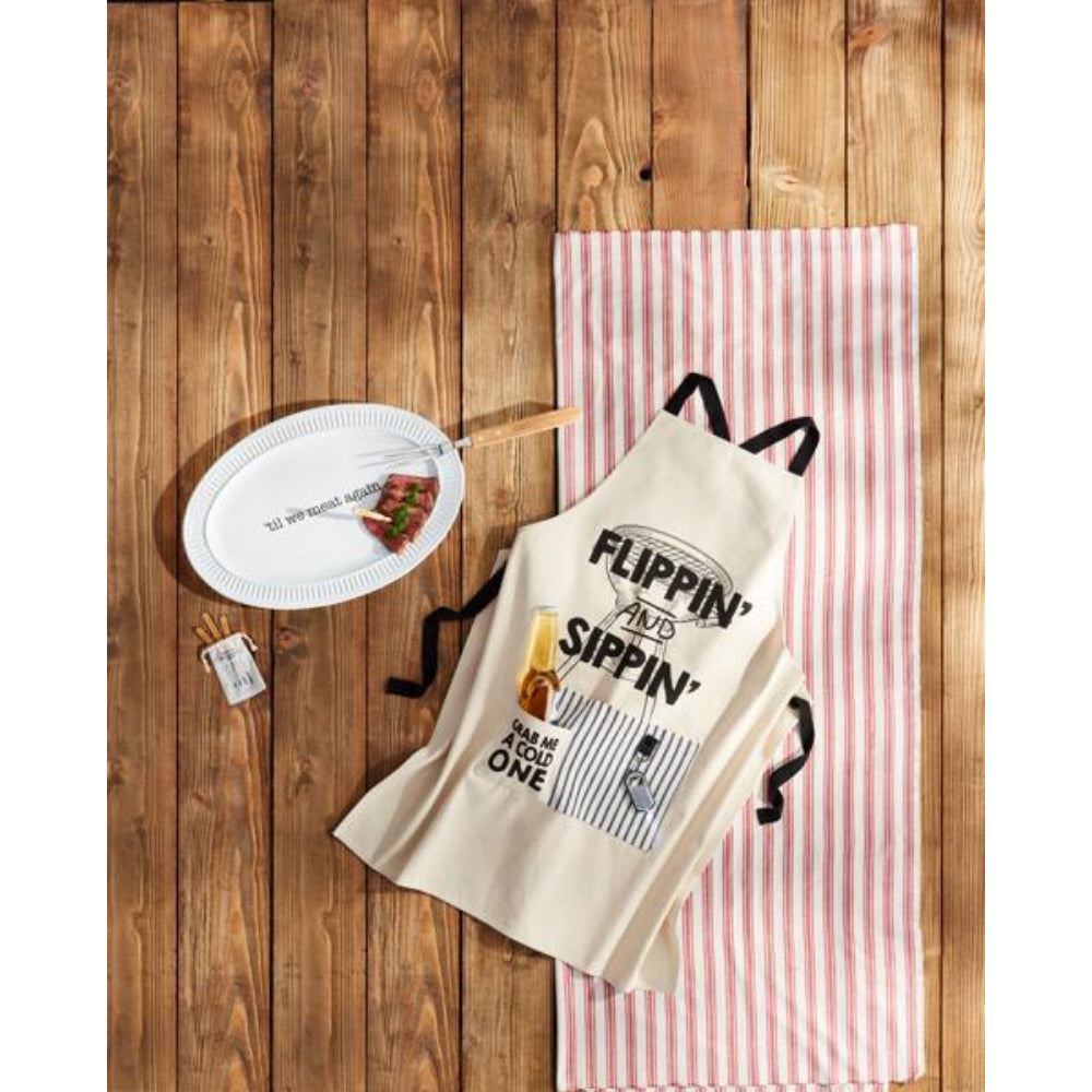 A flat-lay of a beige canvas apron with black straps and a 'FLIPPIN' AND SIPPIN'' graphic, styled on a rustic wooden background next to a pink and white striped towel, a paper plate, and a small matchbox.