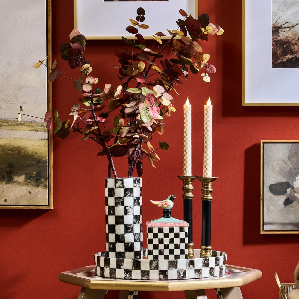 Decorative items including a vase with flowers, candles, and checkered containers on a table against a red wall with framed art.
