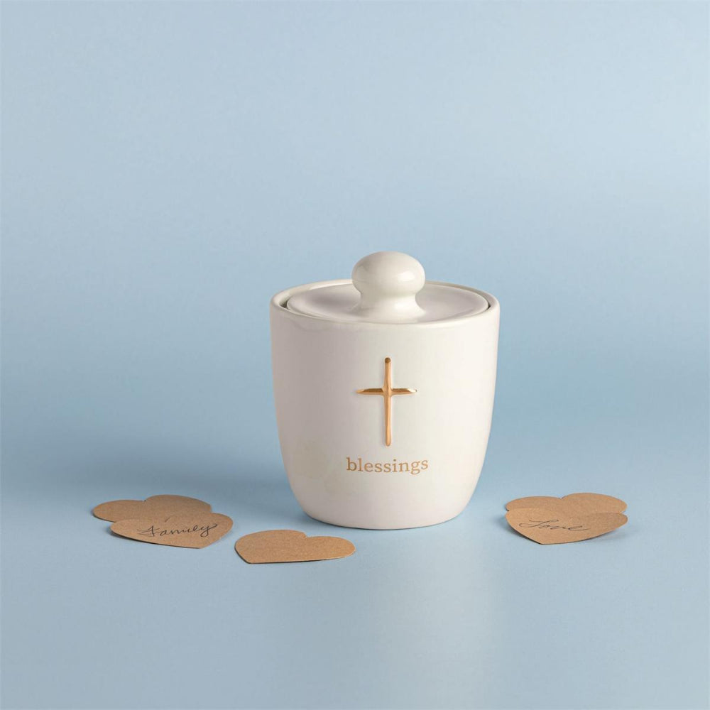 A cream-colored ceramic lidded jar with a gold cross and the word 'blessings' printed on the front, set against a light blue background with three small brown heart-shaped cards.