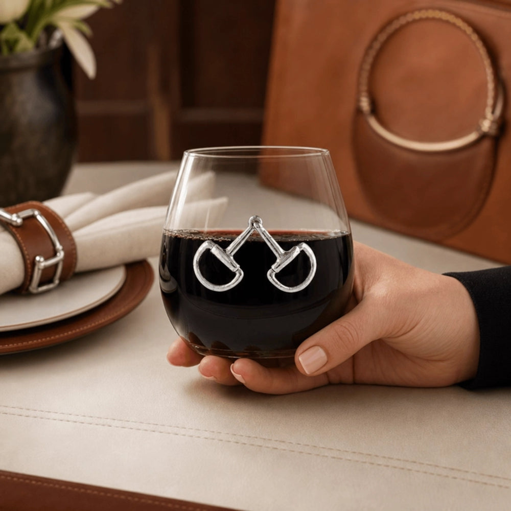 A hand holding a stemless wine glass with a silver horseshoe design on the side, filled with red wine, set in a warm, rustic dining environment with leather and wood tones.