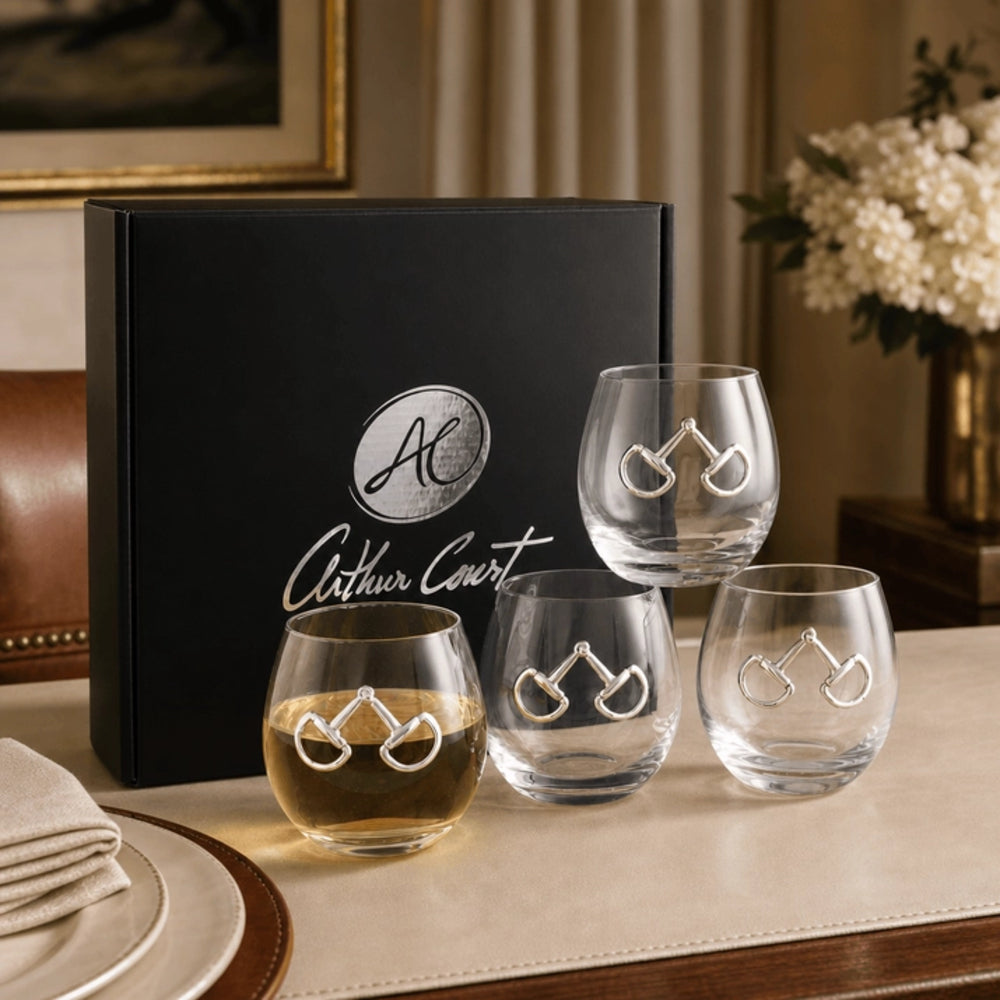 Four clear stemless wine glasses with silver horse-bit engravings arranged on a table in front of a black Arthur Court gift box. The setting includes a beige tablecloth, a plate, folded napkin, and a vase of white flowers in a warm indoor environment.