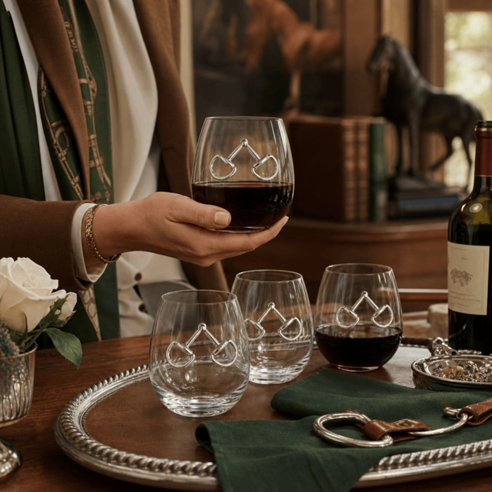A person holds a clear stemless wine glass with an engraved geometric logo, filled with red wine, in a warm, rustic indoor setting with other matching glasses on a silver tray.