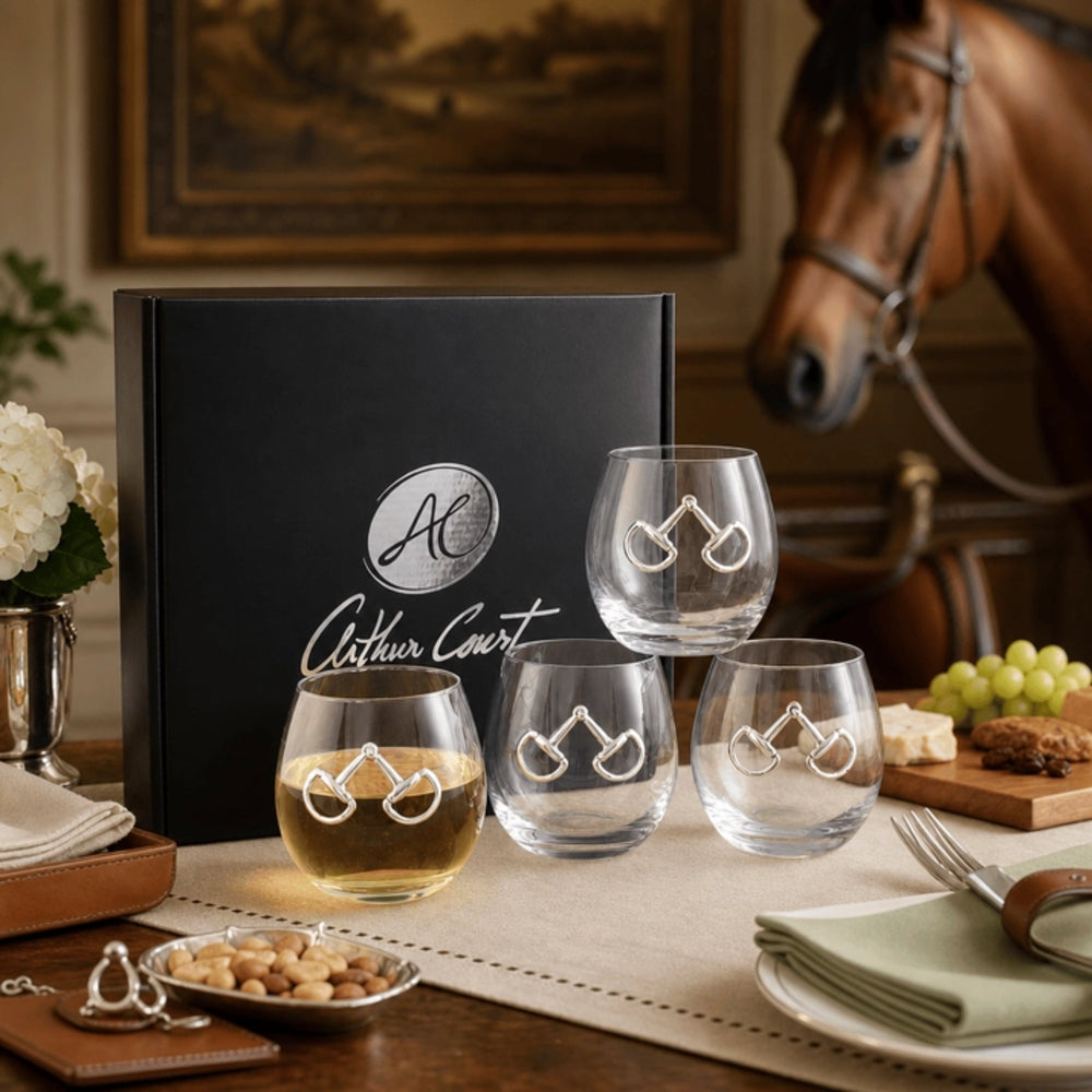Four clear stemless wine glasses with a silver horse-bit design on the front, displayed in front of a black Arthur Court gift box on a dining table. A horse head model and a horse figurine are visible in the background, along with food and table setting details.