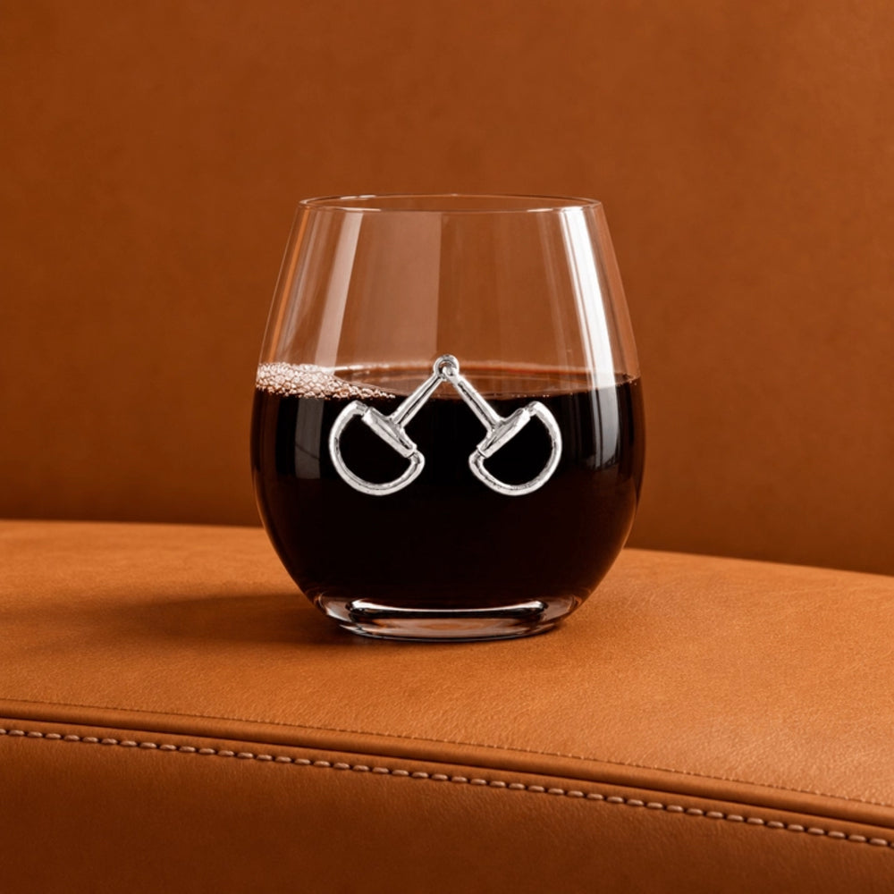 A clear stemless wine glass filled with dark red wine, featuring a silver metallic horsebit design on the front, resting on a brown leather surface against a matching brown background.