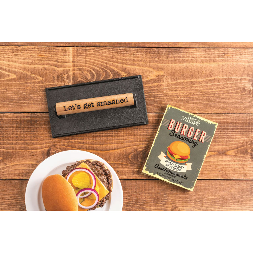 A burger on a plate with seasoning and "Lets get smashed" grill press on a wooden surface.