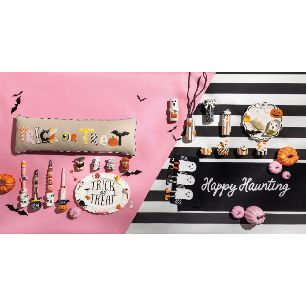 A Halloween-themed tabletop display featuring a pink and black striped table runner with 'Happy Haunting' in white script, surrounded by small witch hats, pumpkins, candy corn, bats, and a 'Trick or Treat' sign. A matching embroidered 'Trick or Treat' pillow sits on a solid pink background.