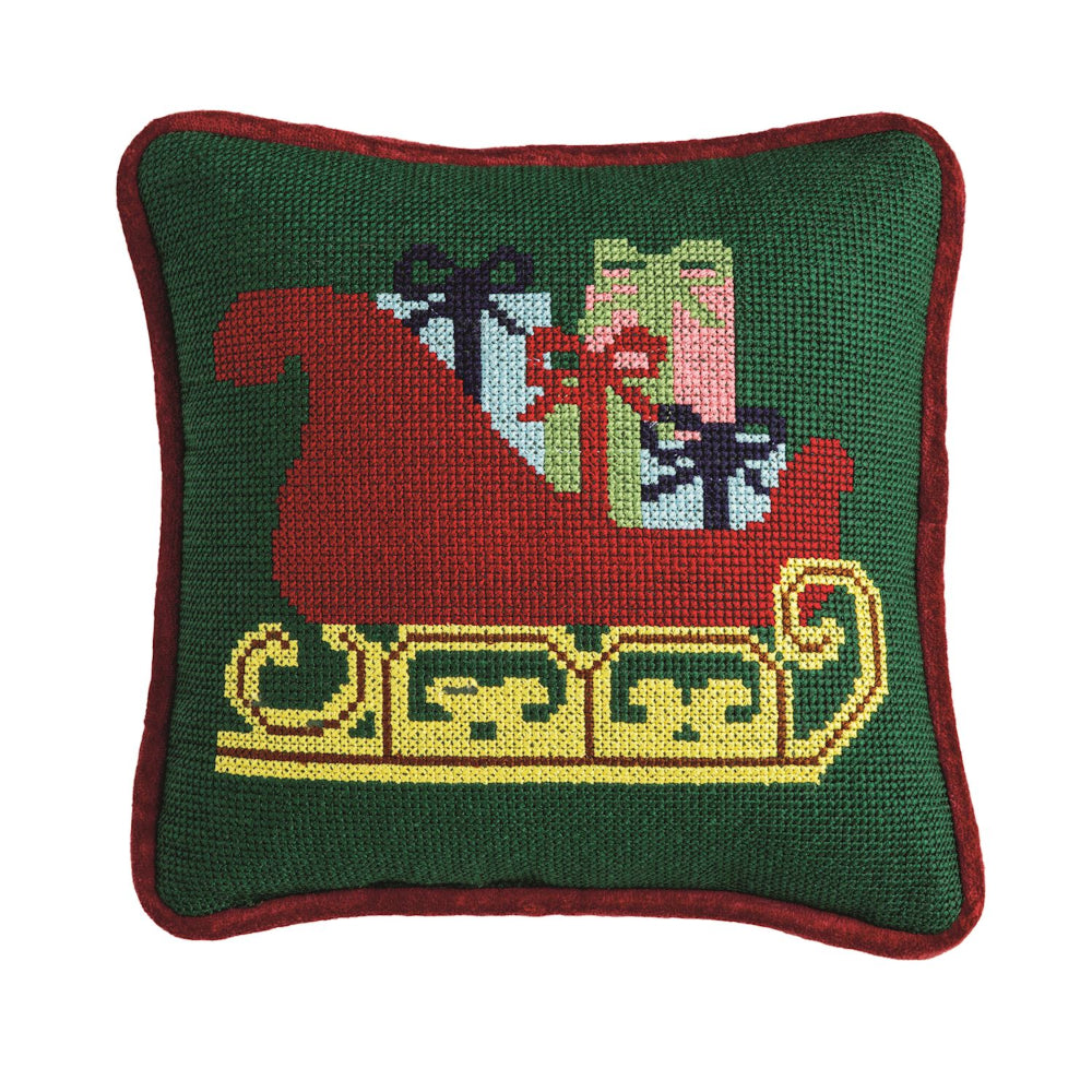 Decorative pillow with a needlepoint design of Santa in a sleigh with presents on a green background.