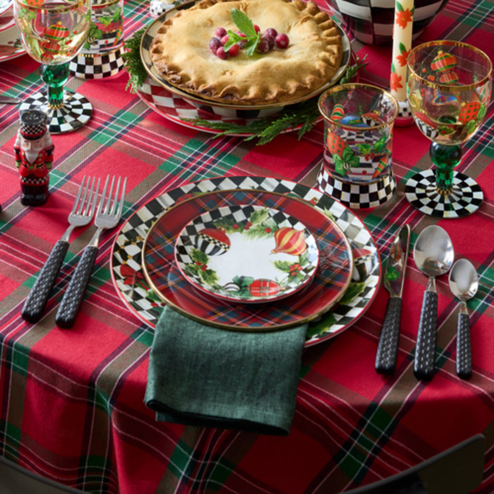 A festive holiday table setting with a red tart on a plaid tablecloth, matching decorative plates, patterned drinking glasses, silverware with black handles, and a folded dark green napkin.