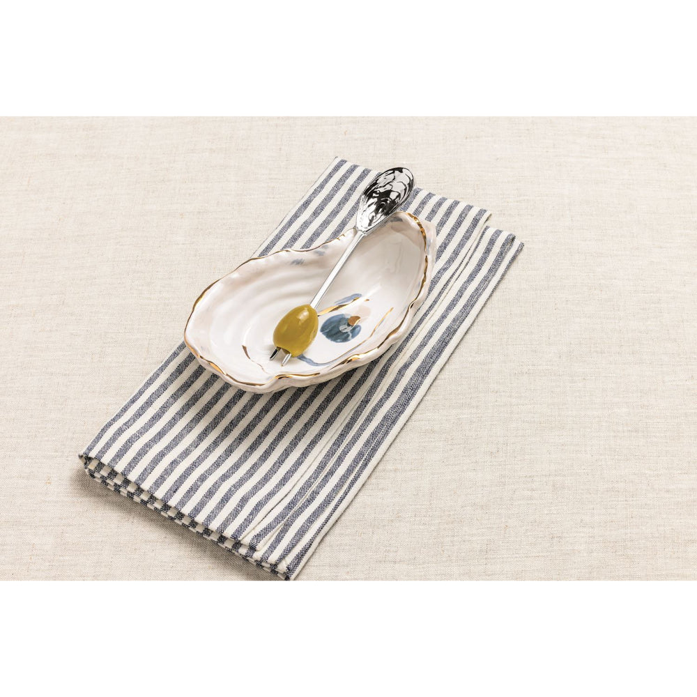 A small oval ceramic dish with a gold-trimmed rim and painted blue floral detail, holding a spoon with a green olivette, placed on a navy-and-beige striped cloth over a light beige textured surface.