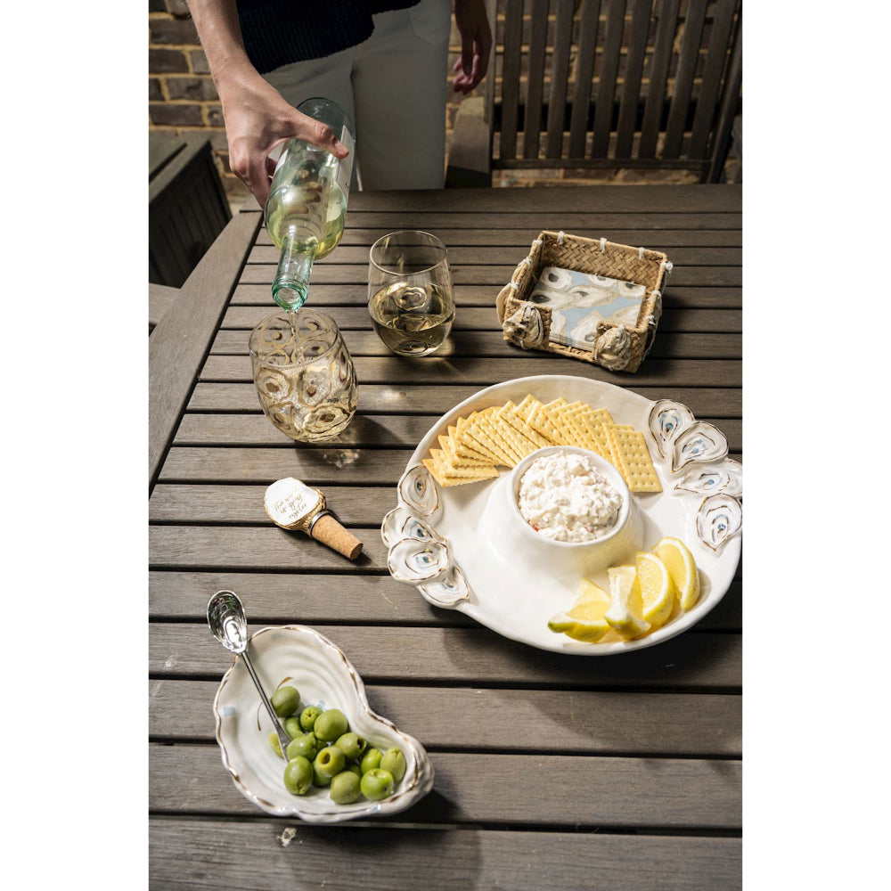 An outdoor dining scene on a dark wooden slat table with a hand pouring white wine from a green bottle into a textured glass. A white serving platter with scalloped edges holds crackers, a bowl of white cheese, lemon wedges, and a heart-shaped dish of green olives.