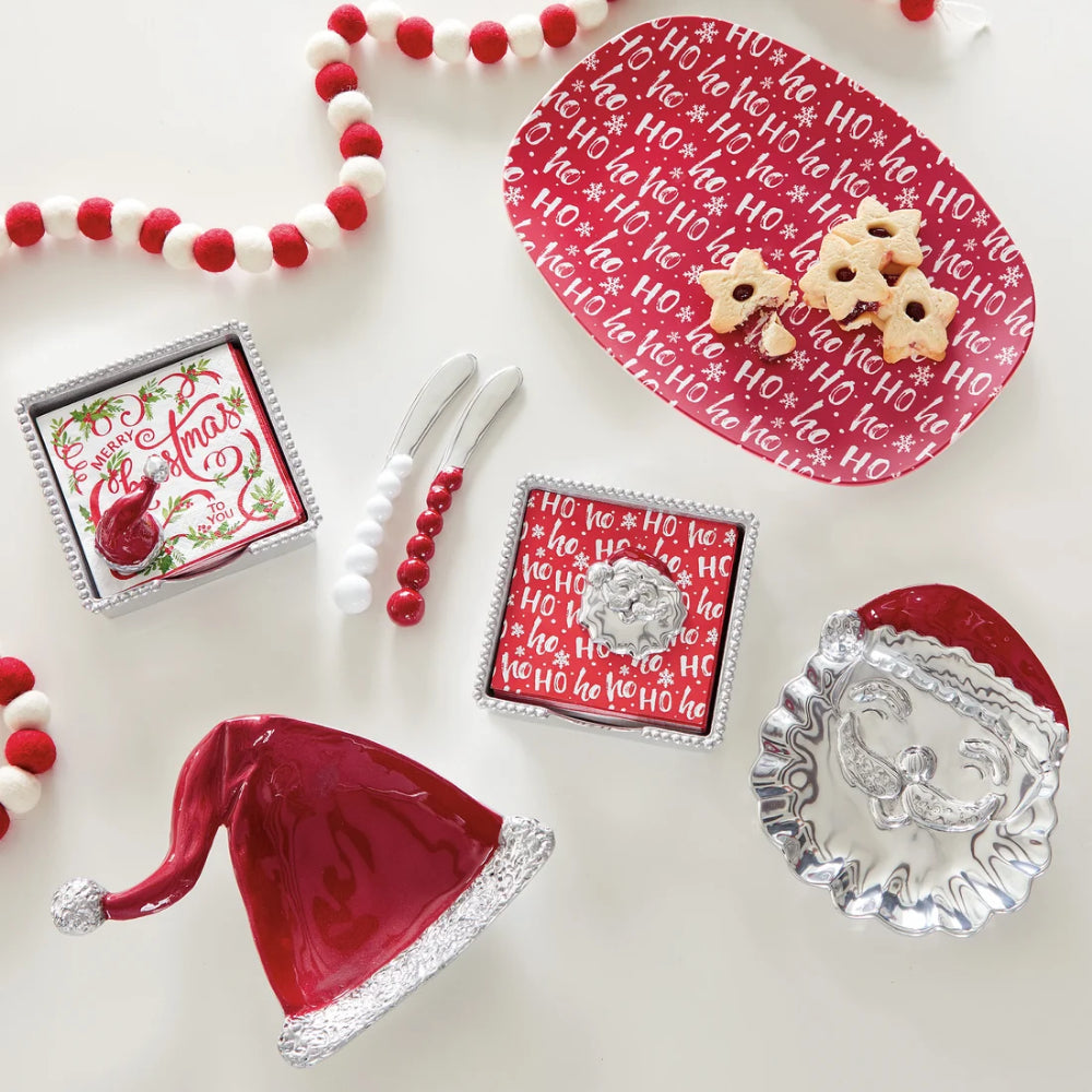 Red santa hat beaded napkin holder with white and red "Merry Christmas" napkins and santa hat weight with other holiday items on a white backround. 