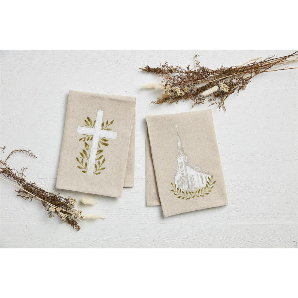 Two beige fabric napkins with embroidered religious designs, one featuring a white cross with olive branches and the other a white church with olive branches, styled on a white wooden surface with dried floral accents.