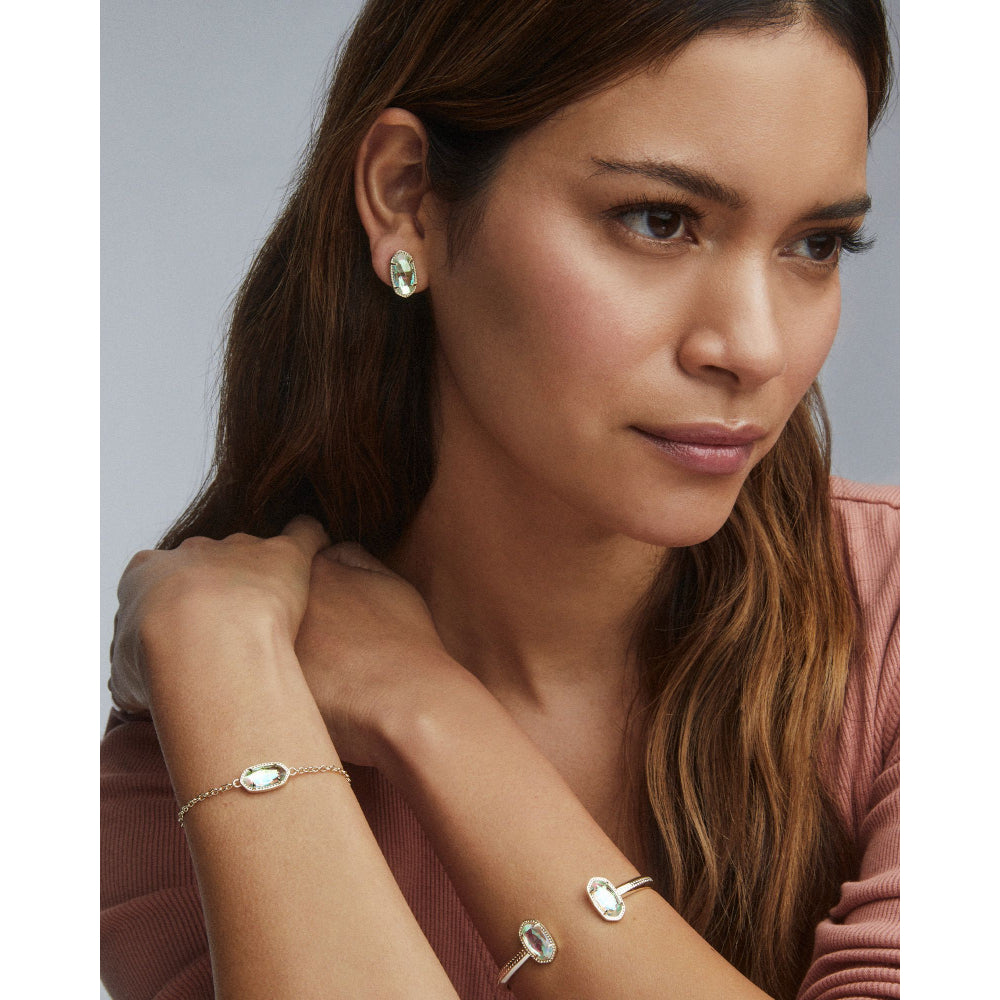 A woman wearing delicate silver-toned jewelry, including a small oval stud earring and matching oval bracelet, paired with a dusty rose top against a neutral gray background.