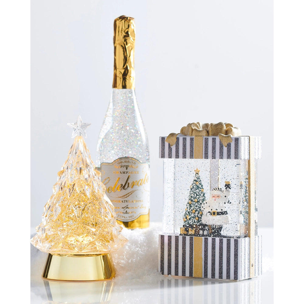 Three holiday-themed party decorations arranged on a white surface against a light gray background: a glittery champagne bottle with a gold foil top, a small illuminated gold Christmas tree on a gold base, and a snow globe in a striped gift box with a gold bow.