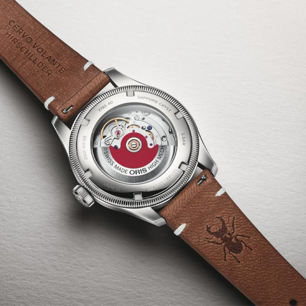 Close-up of the back of an Oris watch with a brown leather strap and a silver-toned case showing the mechanical movement through a transparent backplate. The dial has a red center, and the strap is engraved with 'CERVOLANTE SCHLEIDER' and has a spider logo.