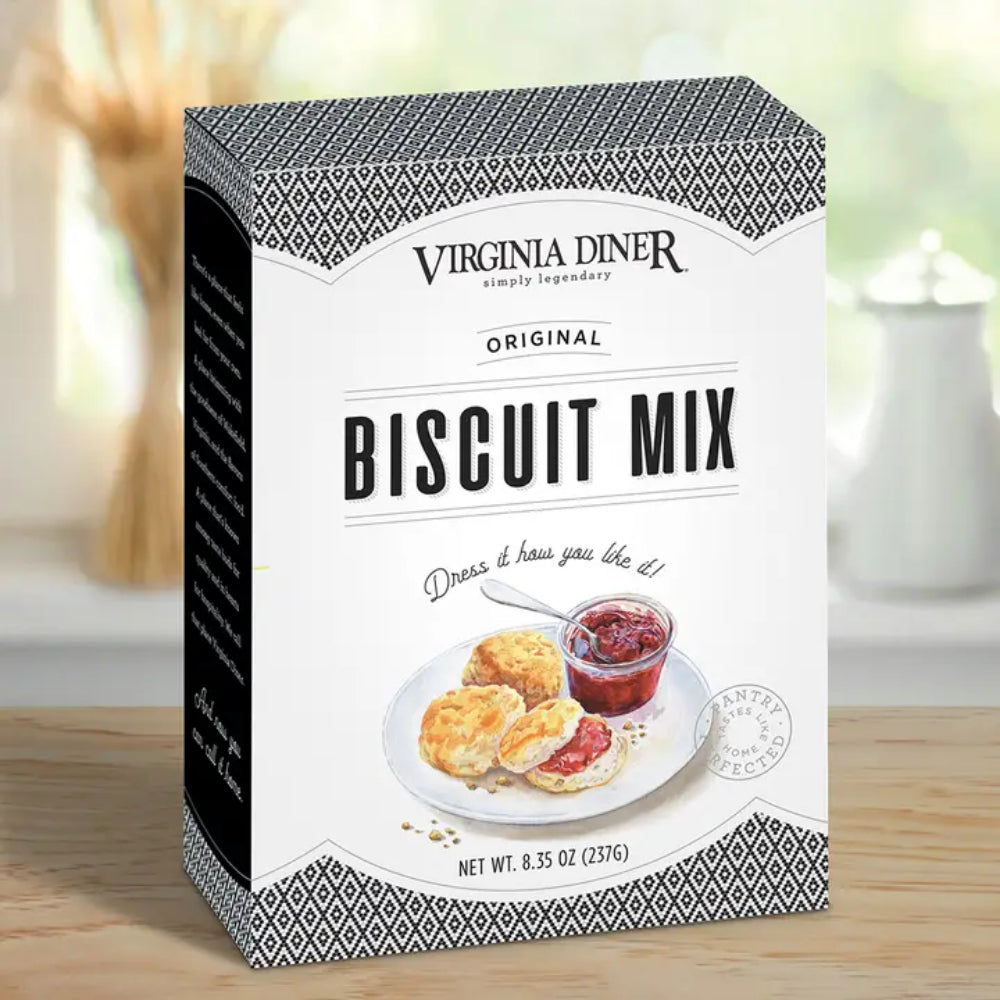 A box of Virginia Diner Original Biscuit Mix standing on a wooden surface with a softly blurred kitchen background. The front of the box is white with black text and a printed illustration of two scone halves with jam and a small bowl of red jam.