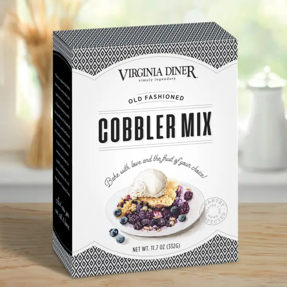 A box of Virginia Diner Old Fashioned Cobbler Mix standing on a wooden surface with a softly blurred kitchen background. The packaging is white with black geometric trim and a photo of a cobbler with an ice cream scoop on the front.