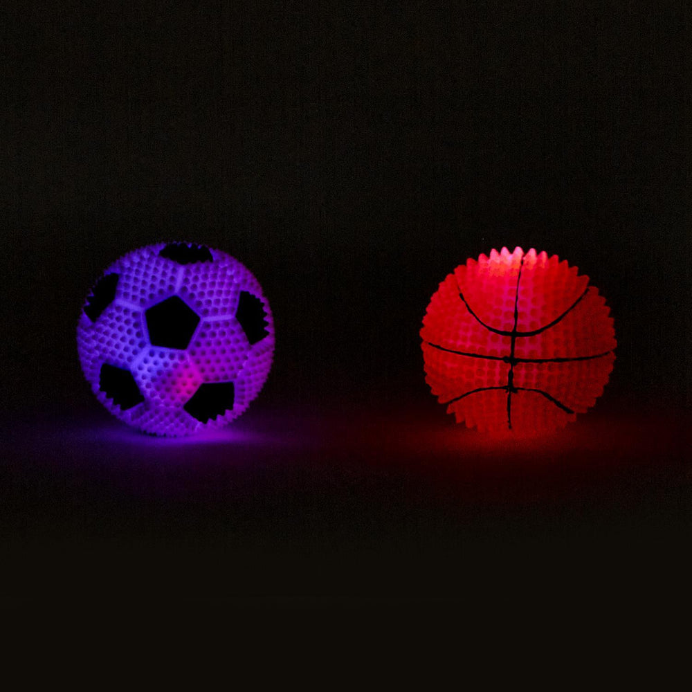 Two textured stress balls, one soccer ball pattern glowing purple and one basketball ball pattern glowing red, resting on a black surface against a dark background.