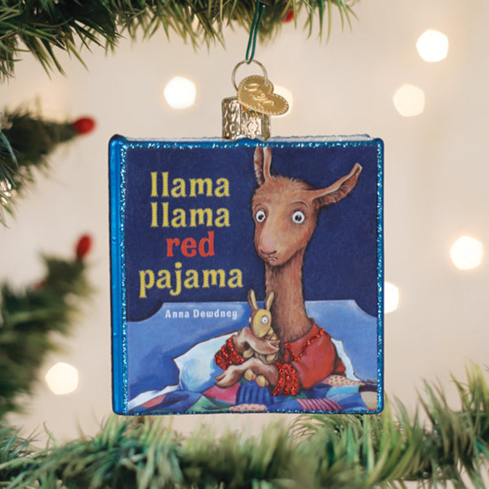 A square Christmas ornament shaped like the children's book cover of 'Llama Llama Red Pajama' hangs from a green Christmas tree branch with warm lights in the background.