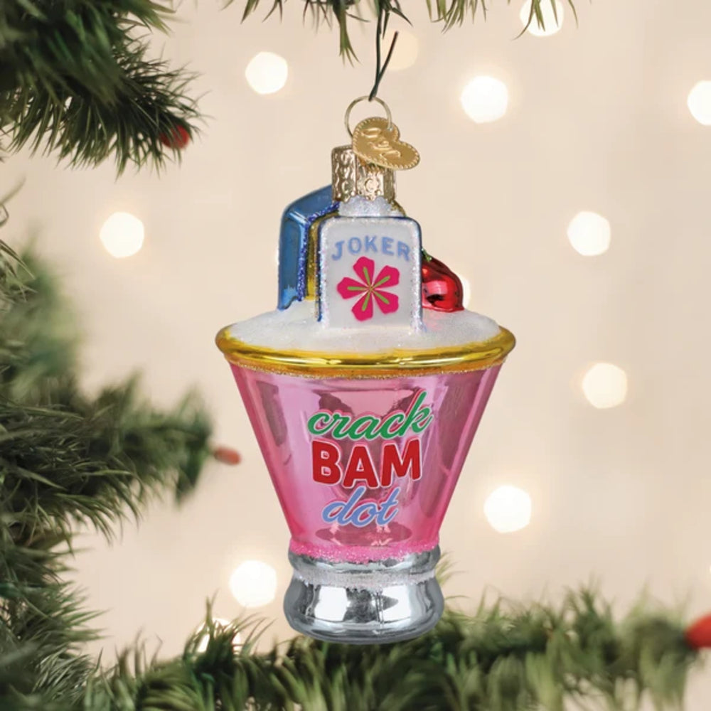A Christmas ornament shaped like a pink cocktail glass with white foam, gold rim, and a small bottle labeled "JOKER" on top. The ornament hangs from a tree branch against a warm, blurred background with bokeh lights.