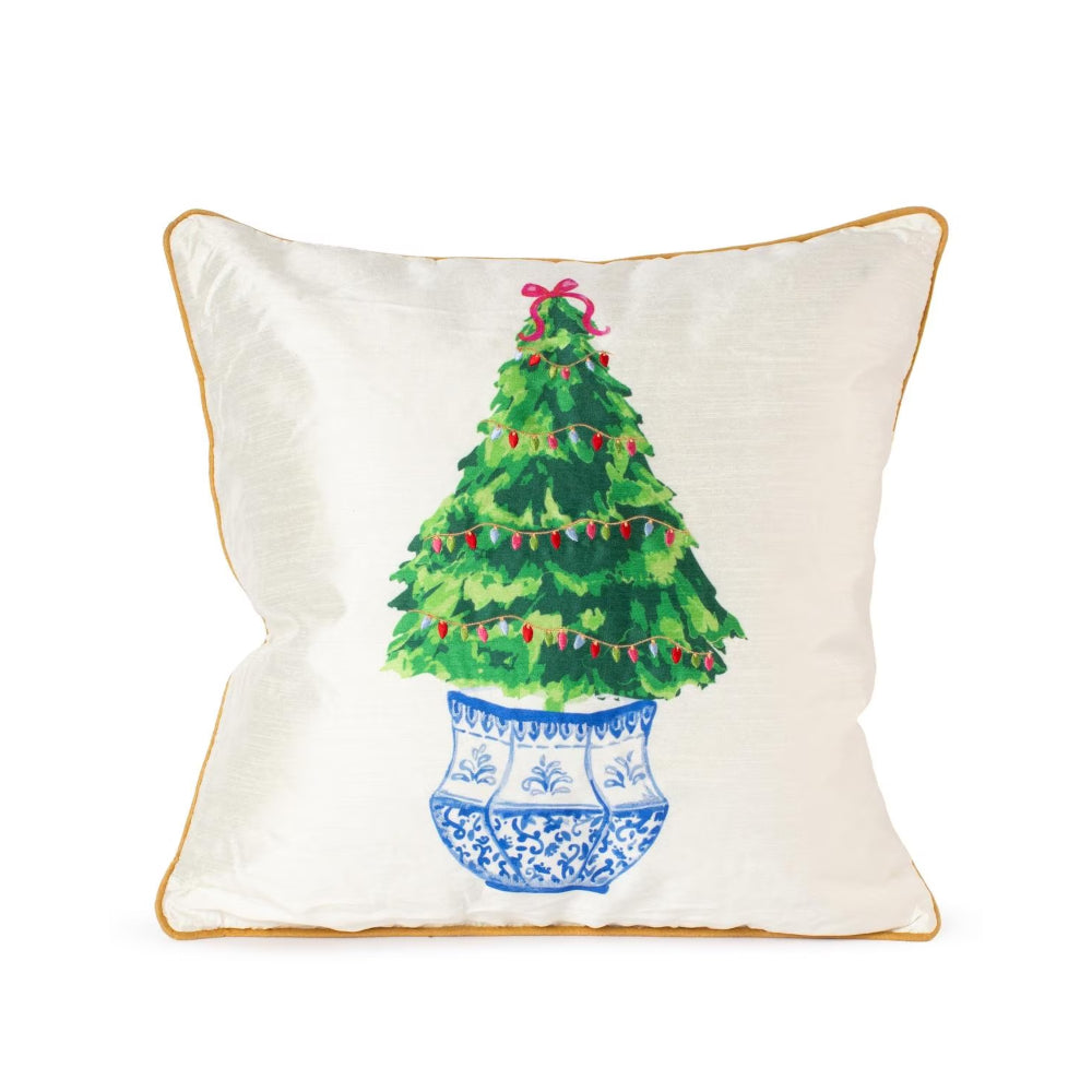 An off-white square throw pillow with a gold-colored piped edge, featuring a printed illustration of a green Christmas tree in a blue patterned pot with red ornaments and a pink bow.