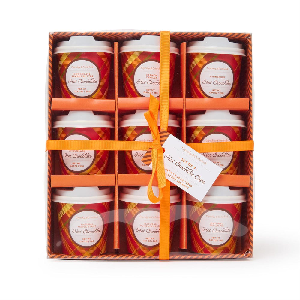 A gift box of nine hot chocolate cups arranged in a 3x3 grid, wrapped with an orange ribbon and a small hanging tag. The cups have red and orange plaid labels with white lids, and the box itself has a brown patterned border.