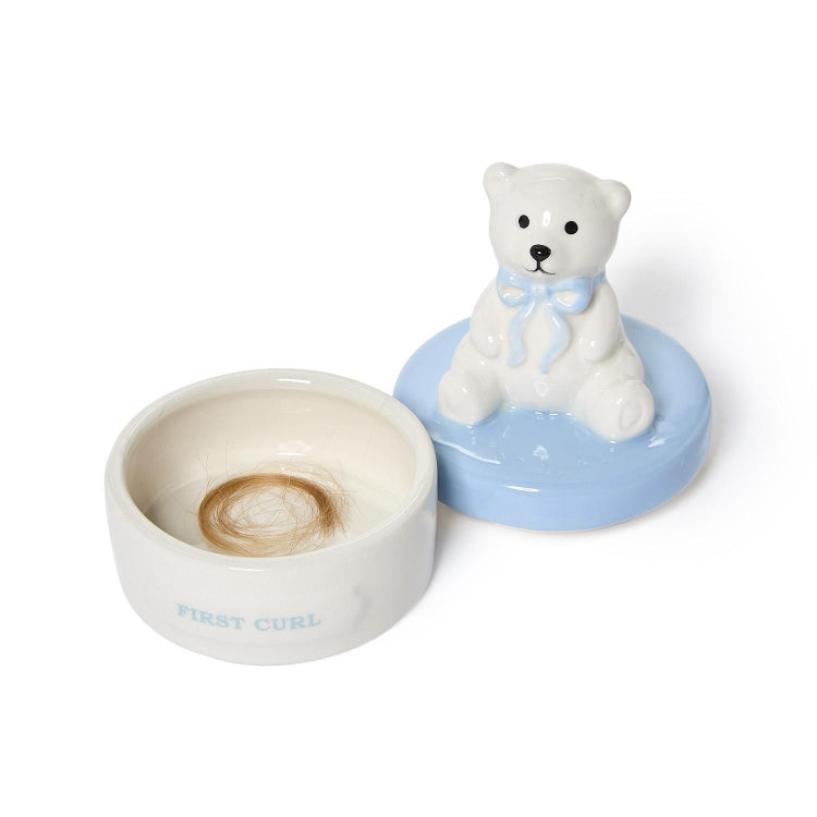 A ceramic hair trap container shaped like a white bear sitting on a light blue base, with the lid off to reveal a small brown hair coil inside a white bowl. The lid has a white bear figurine and the bowl is printed with the text 'FIRST CURL' in light blue.