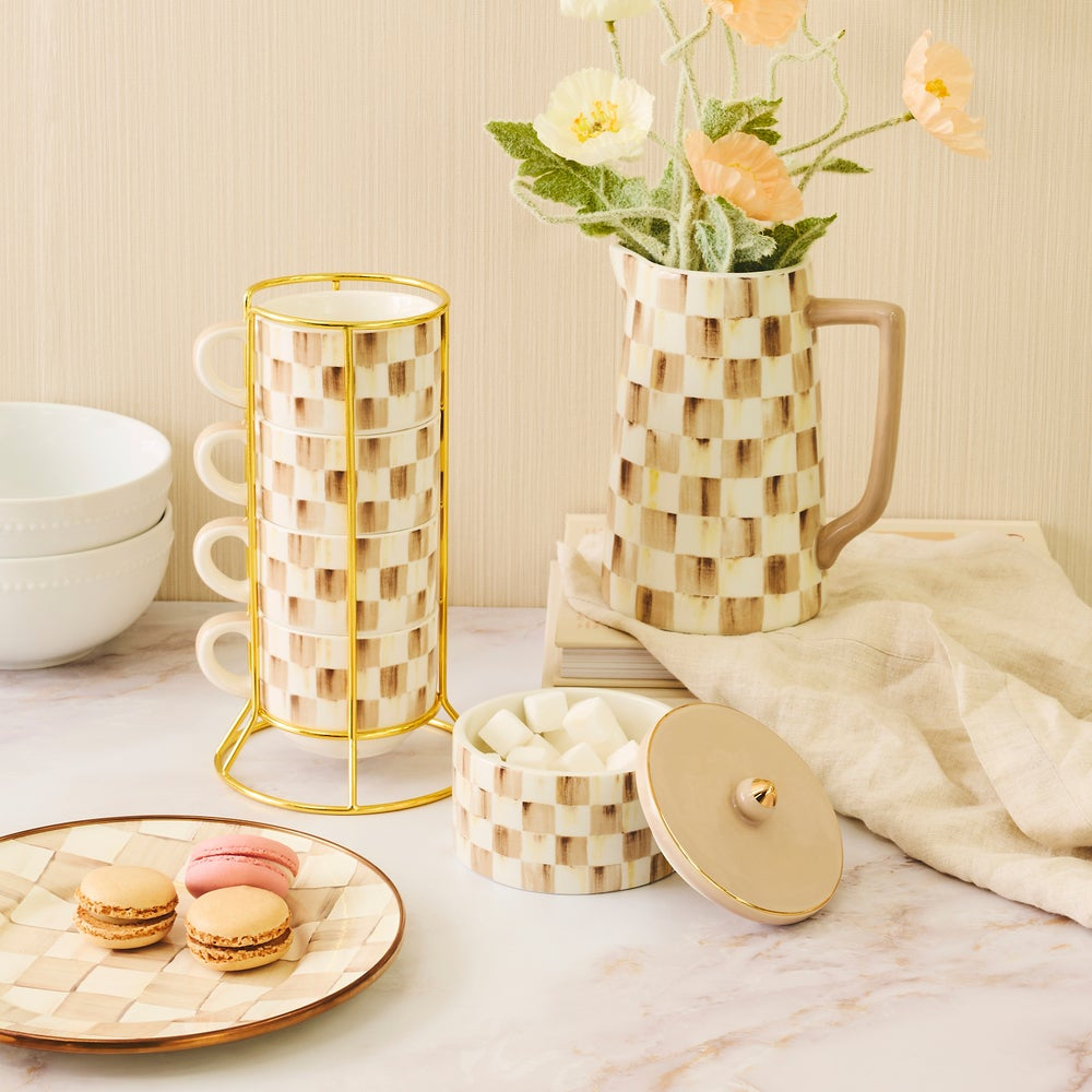 A coordinated set of cream and tan woven ceramic tableware with gold-tone metal accents, including a pitcher with flowers, a cylindrical mug holder, a small lidded container, and a plate with macarons, styled on a marble surface against a beige wall.