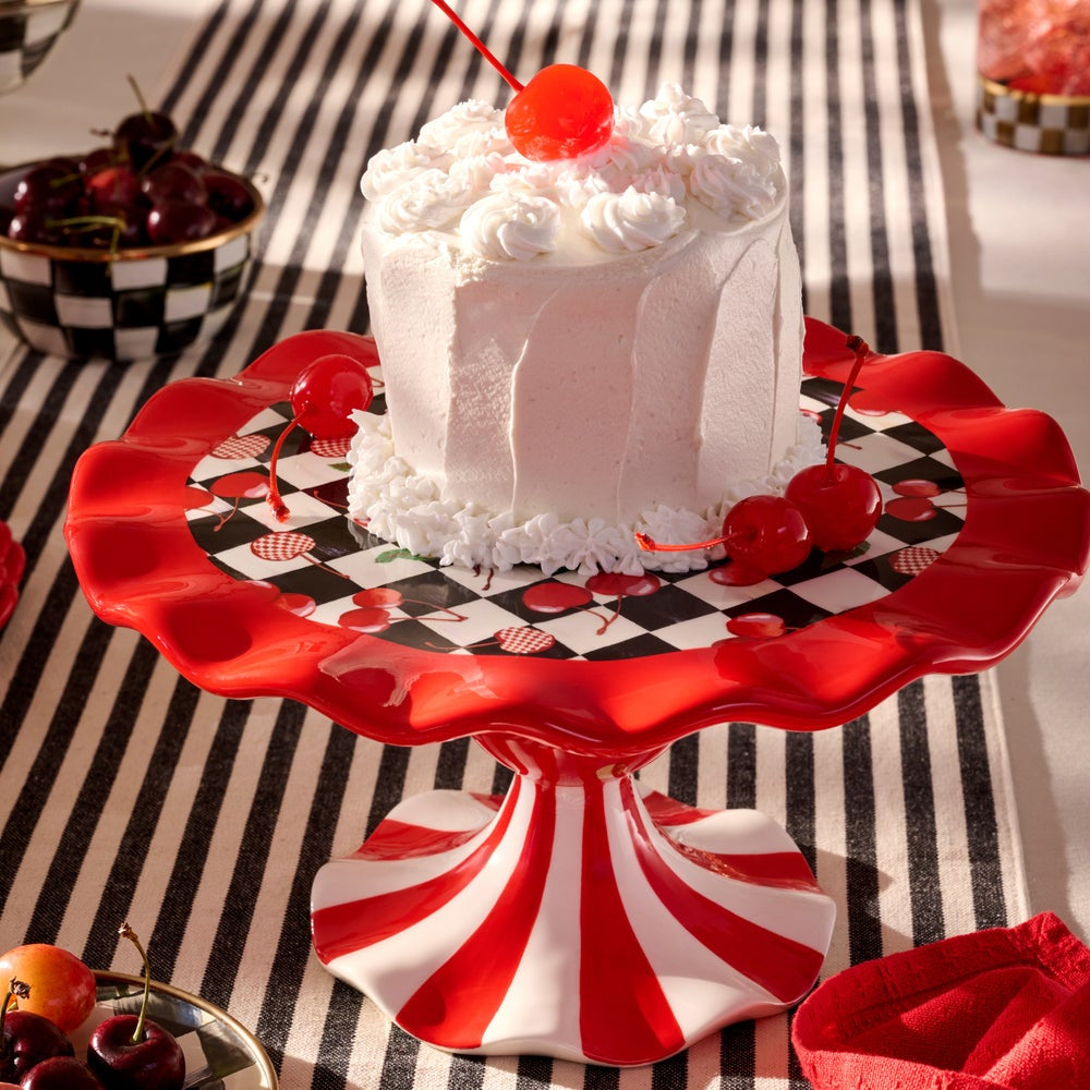 A white frosted cake topped with a bright red maraschino cherry sits on a red-and-white pedestal cake stand with a black-and-white checkered base. The stand is placed on a black-and-white striped tablecloth, with cherries and bowls in the background.