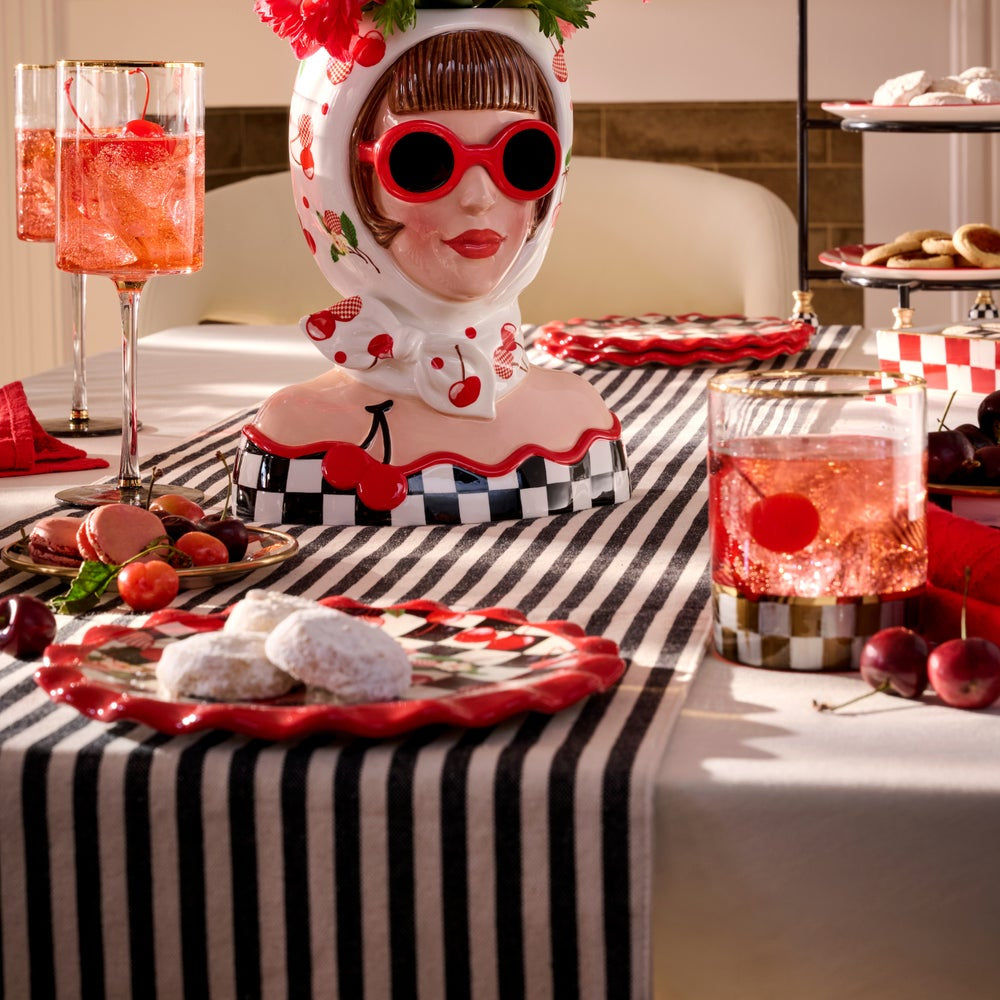 A decorative bust-shaped ceramic container styled on a black-and-white striped table runner, surrounded by red and white party tableware, glasses of pink drink, and pastries in an indoor setting.