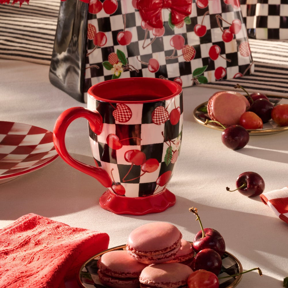 A ceramic mug with a red handle, red base, and a black-and-white checkered pattern decorated with red cherries, styled on a table with macarons, cherries, and a matching gift bag.