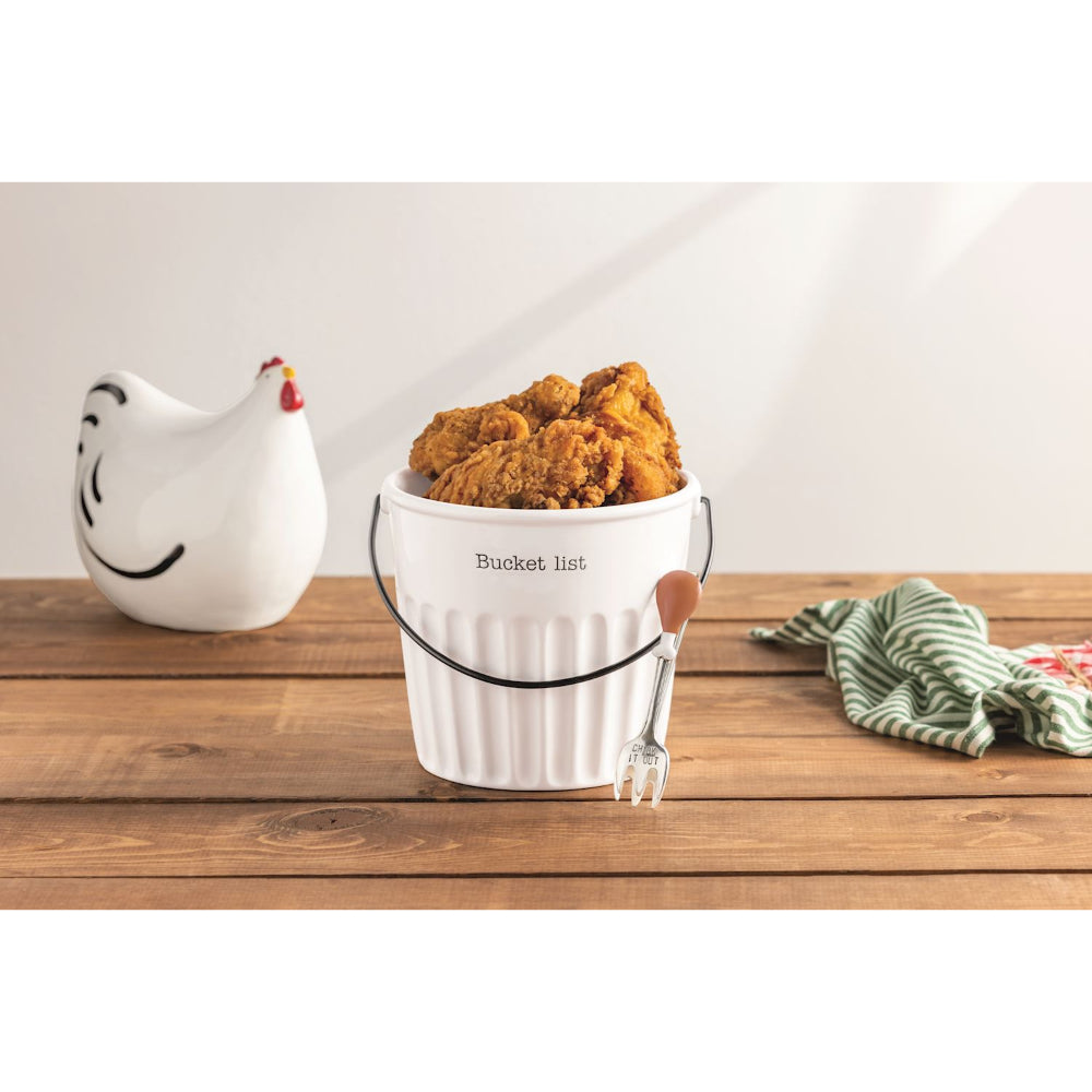 White bucket labeled 'Bucket list' with fried chicken on a wooden surface, next to a ceramic chicken and striped towel.