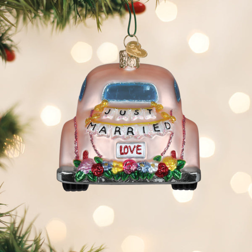 A decorative Christmas ornament shaped like the back of a pink car, hanging from a green string. The ornament has a glossy finish and is decorated with a blue window, a gold bell, a banner reading 'JUST MARRIED' with a 'LOVE' sign, and colorful floral accents at the bottom.