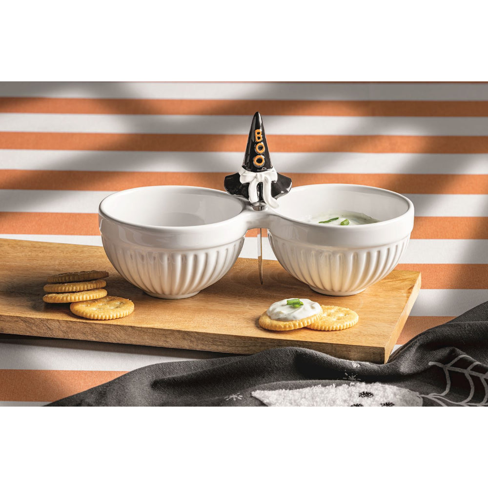 A white ceramic double bowl serving dish with a witch hat topper, styled on a wooden board with crackers against a striped background.