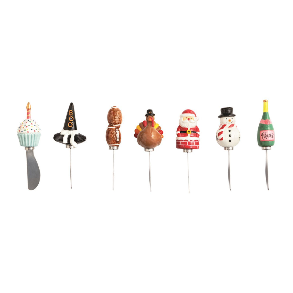 Seven holiday novelty spreaders with festive handles, including a cupcake, witch hat, gourd, turkey, Santa, snowman, and champagne, arranged in a row on a white background.