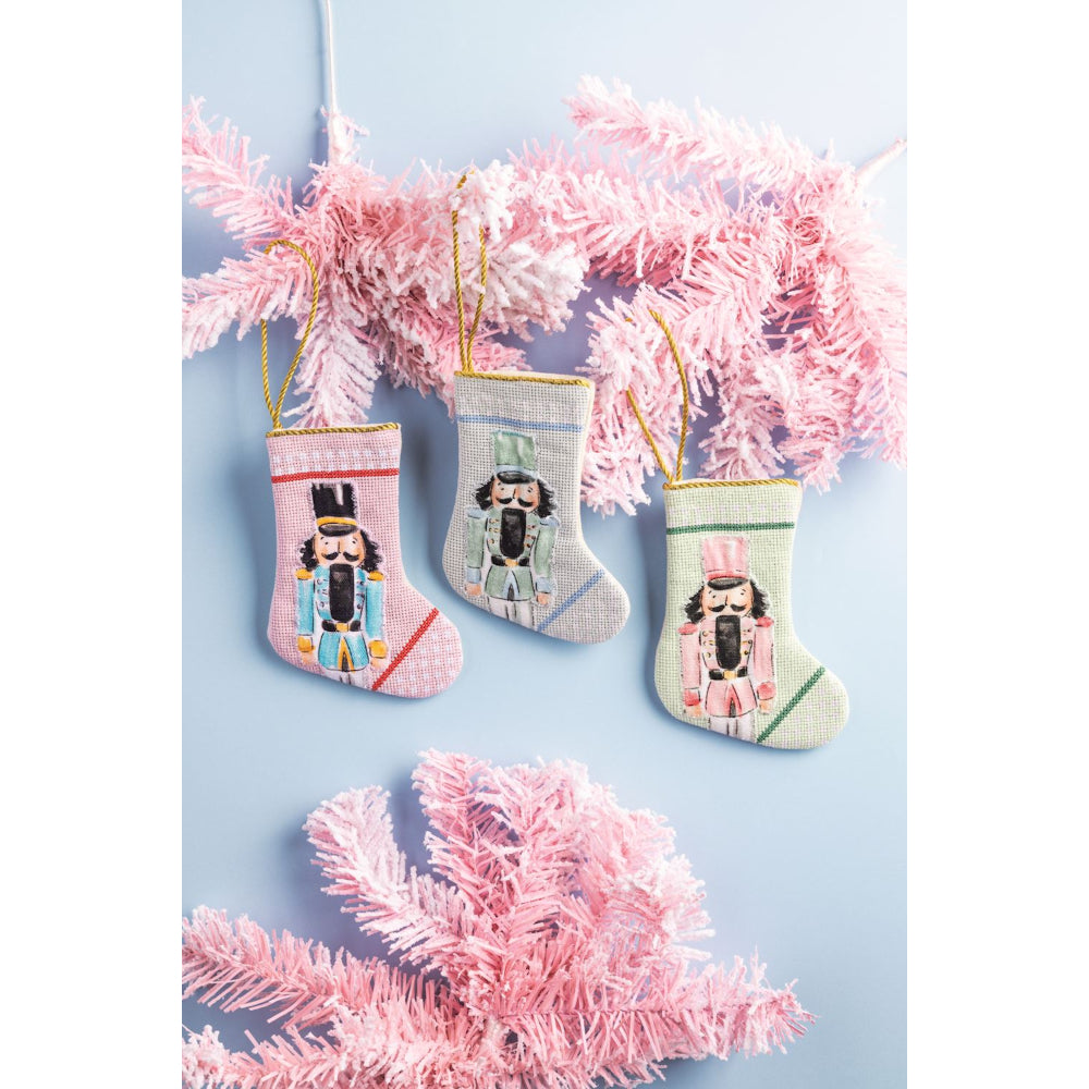 Three small Christmas stockings with nutcracker designs hanging on pink tinsel against a light blue background.