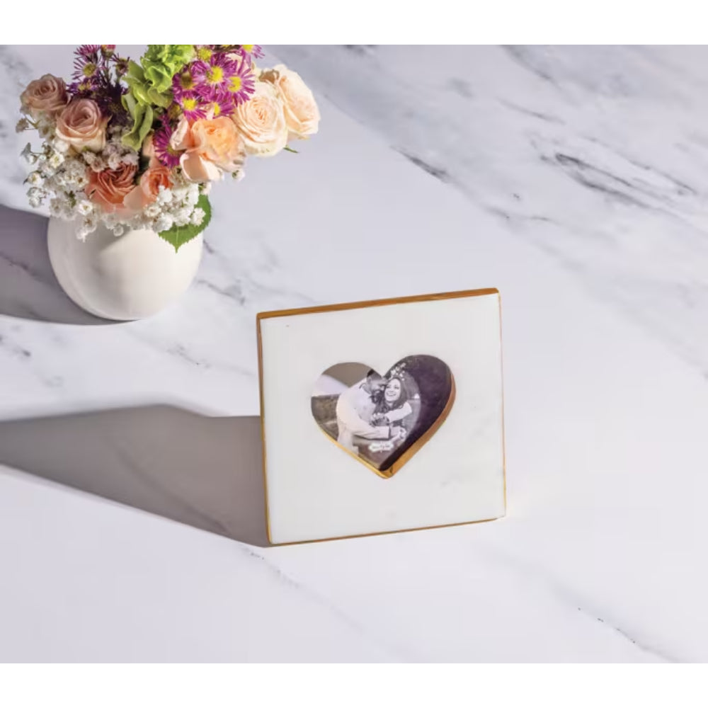 A square white photo frame with a gold border and a heart-shaped cutout in the center, displayed on a white marble surface next to a white vase of flowers.